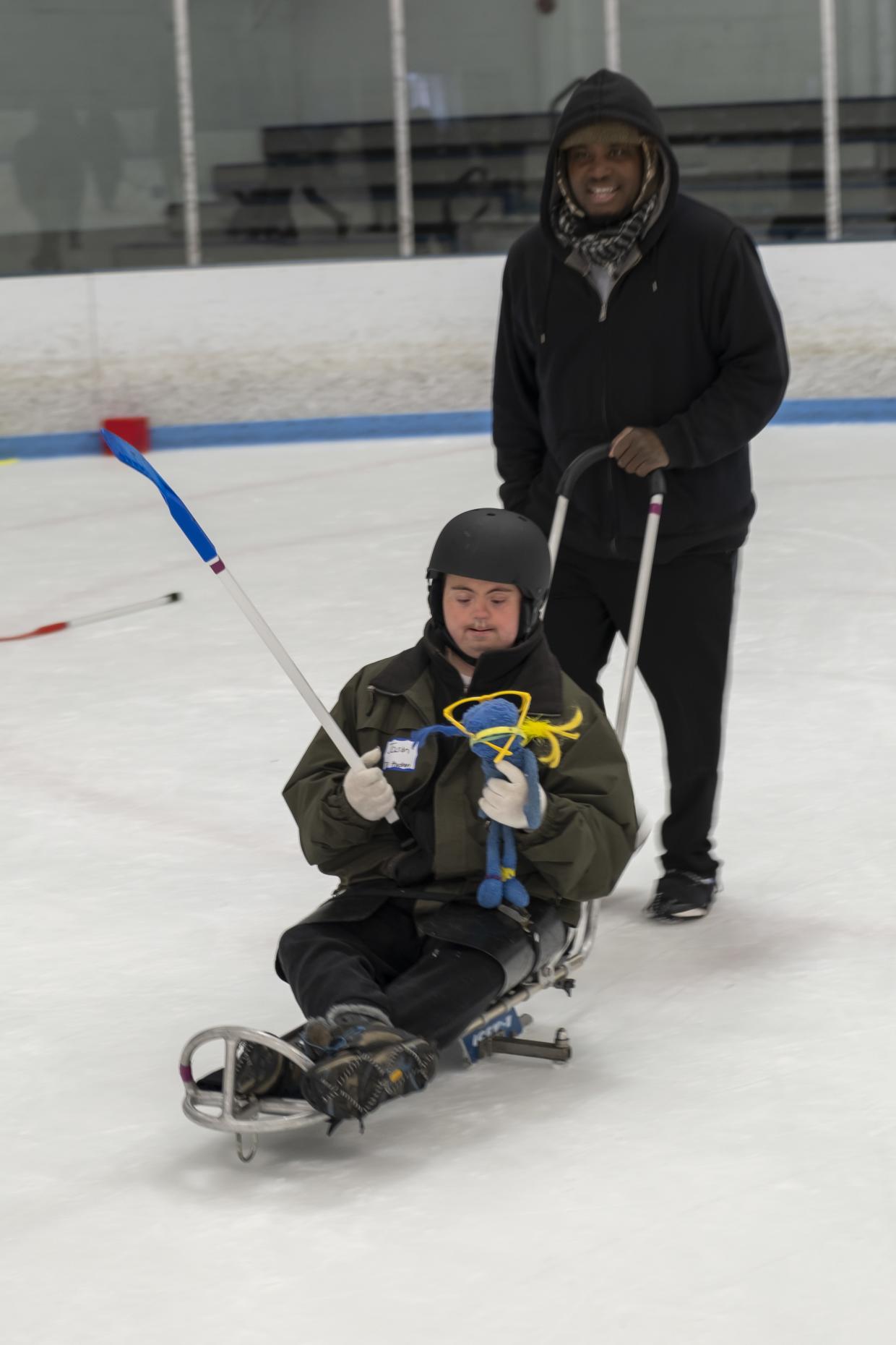 A person in an ice sled is holding a hockey stick and a doll while another person pushes them.