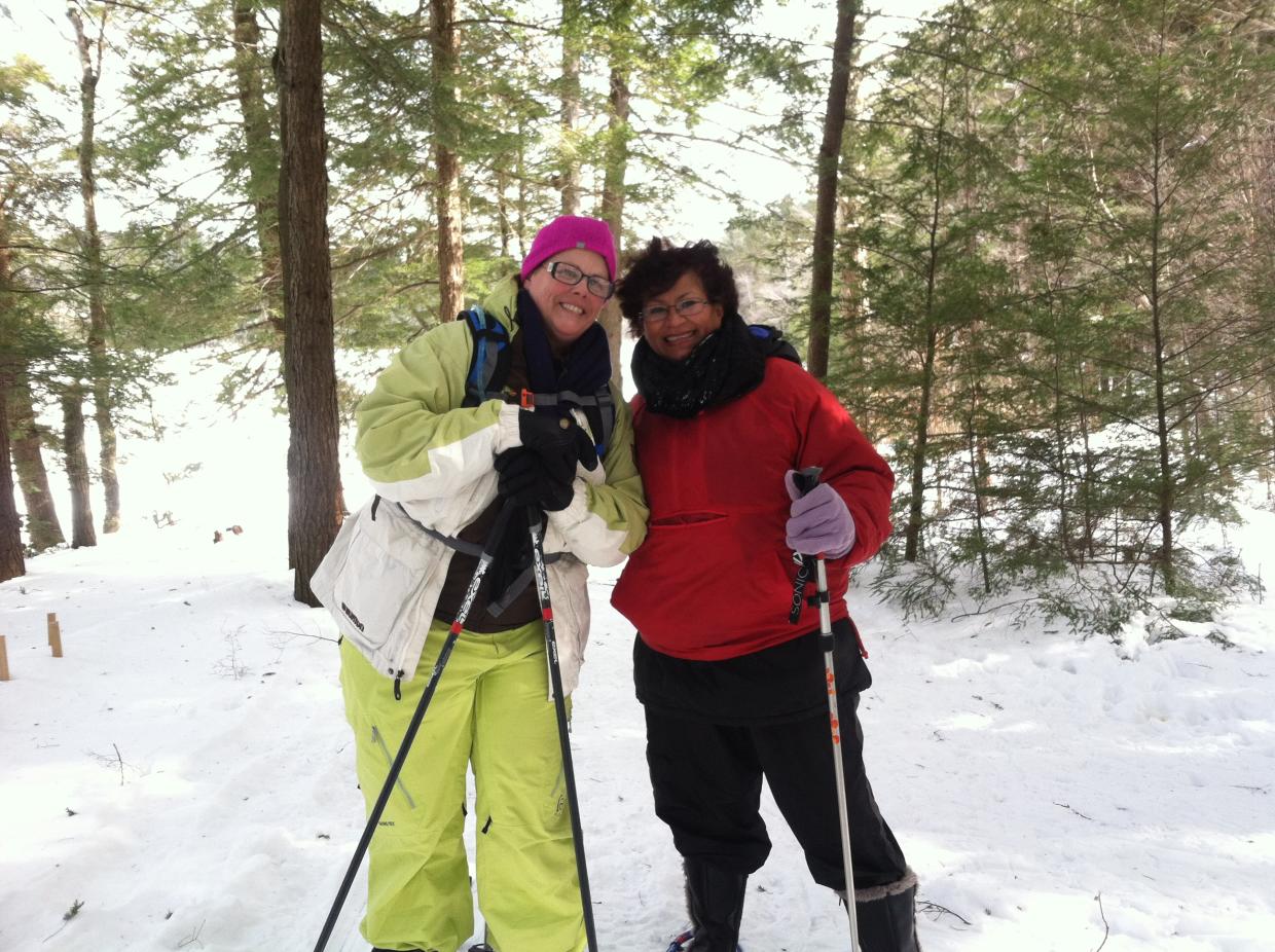 Two women using snowshoes and ski poles in the winter woods smile at the camera.