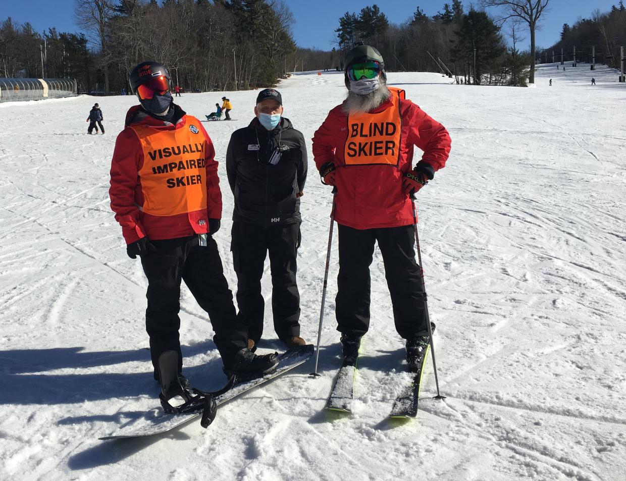 Three skiers stand at the bottom of a snowy hill on a sunny day. One skier has a vest that says visually impaired skier. Another has a vest that says blind skier.