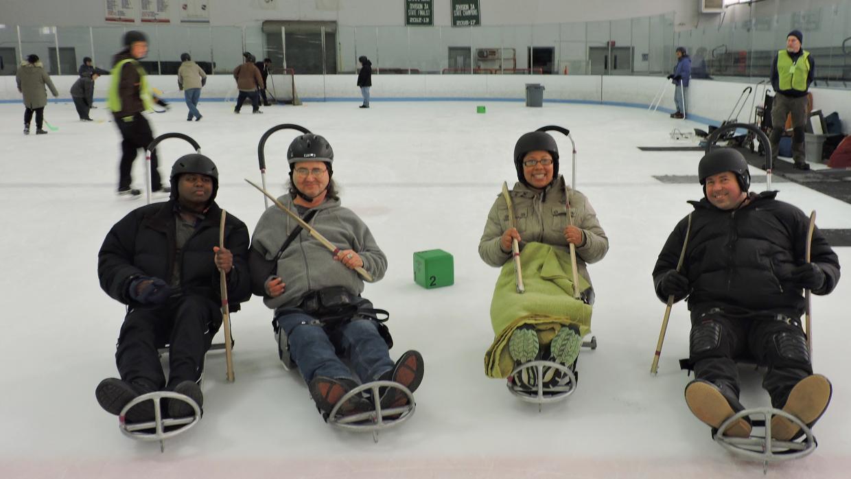 Four sled skaters wearing helmets smile for the camera. behind them, other skaters shoot pucks at a hockey net.