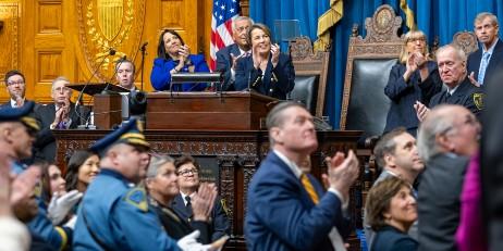 Governor Maura Healey and Lt. Gov Kim Driscoll and a crowd of people in the State House, all looking up and to the right and applauding