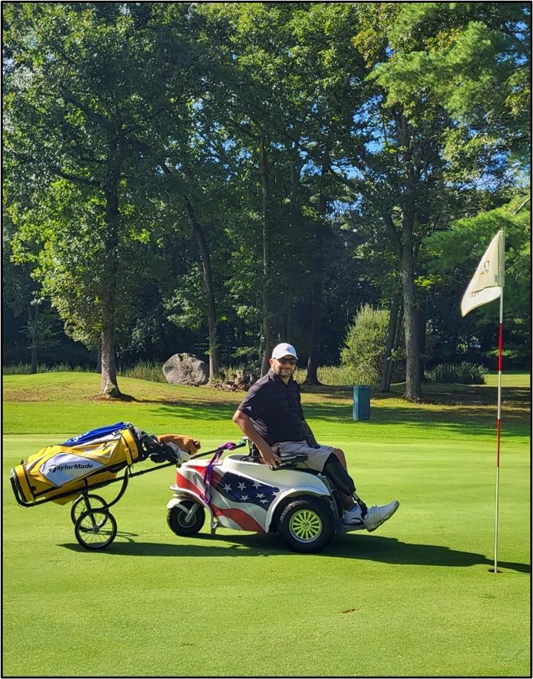 An amputee is sitting on a paragolf wheelchair. Their golf club bag is being pulled from hind on a wheeled cart. They are on green grass near a golf hole marked by a flag.  It is a sunny day and trees are in the background.