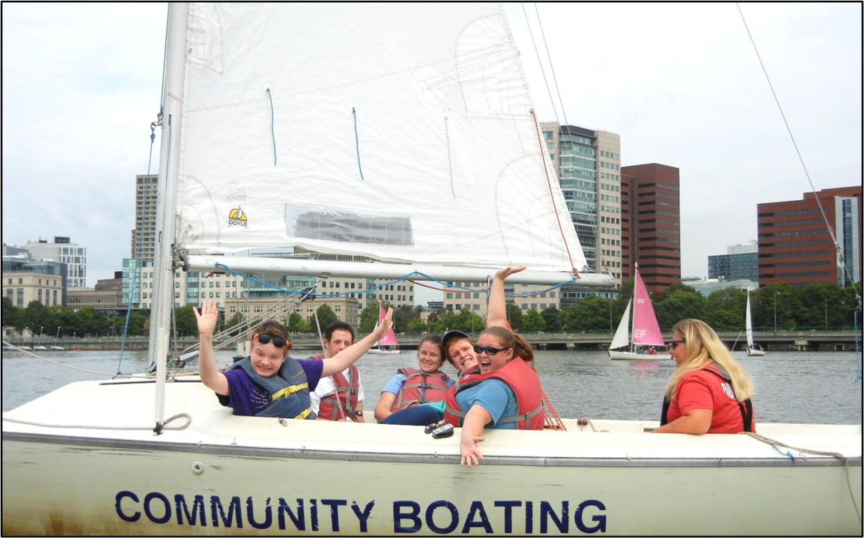 A group of people wearing life vests are sitting in a sailboat  smiling and waiving. The sail boat is on a river, has a single white sale and is there is a Community Boating sign on the side. There are tall city buildings and other sail boats in the background.