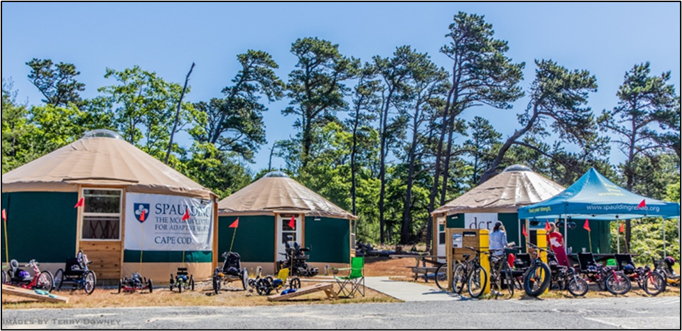 Many styles and sizes of adaptive bicycles are parked in front of 3 accessible yurts and a Spaulding pop up canopy.  There is a Spaulding banner on one yurt and a DCR banner on another yurt.  It is a sunny day and there are green pine trees in the background.