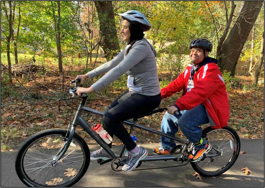 Two people wearing helmets are riding a tandem upright bicycle on a tree-lined path.   