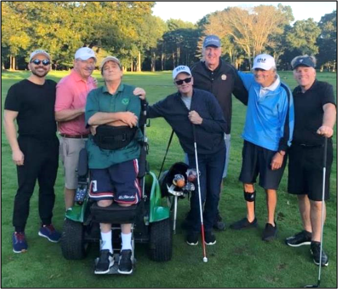 A group of people are standing on a green grassy golf course posing for a picture. One person is standing using ParaGolfer and one person is using a white cane.