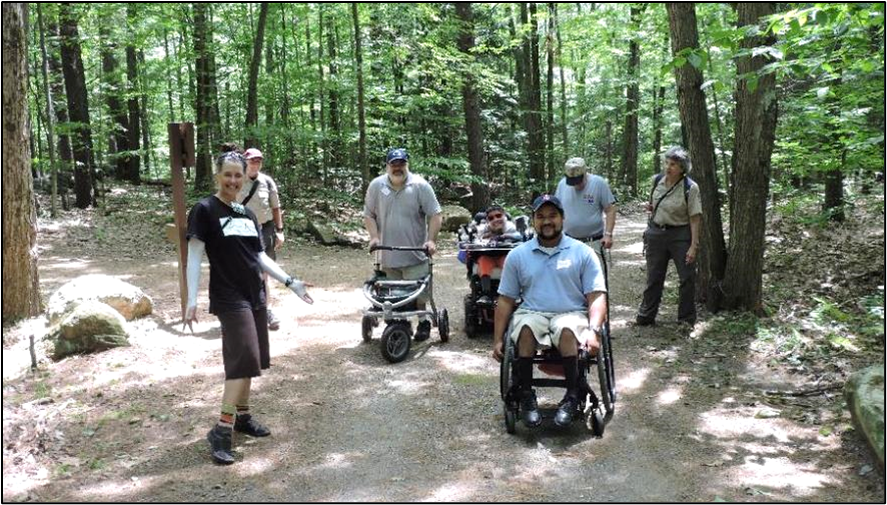 A group of people using a variety of mobility devices on a trail in the woods.
