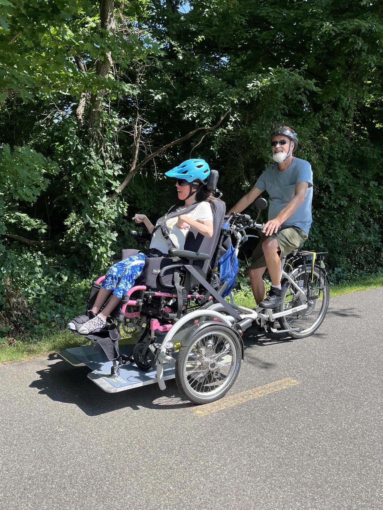 Two people are riding a wheelchair tandem trike on a paved path. 