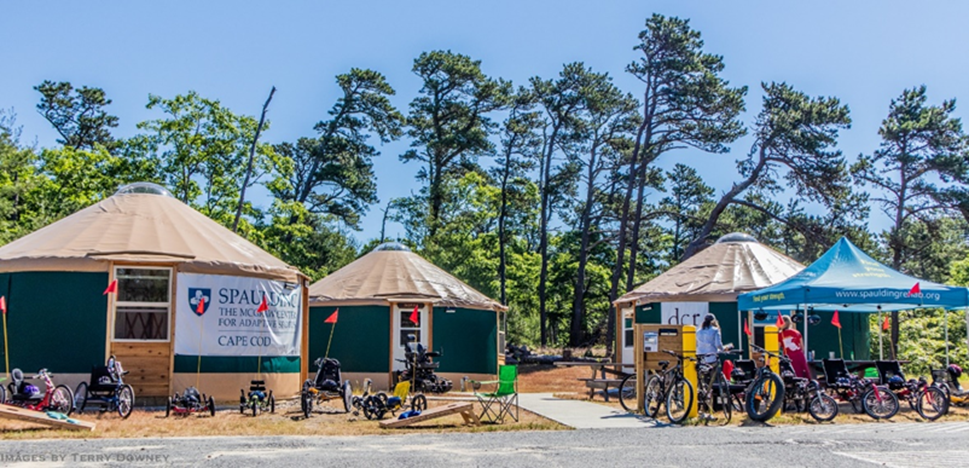Many styles and sizes of adaptive bicycles are parked in front of 3 accessible yurts and a Spaulding pop up canopy.  