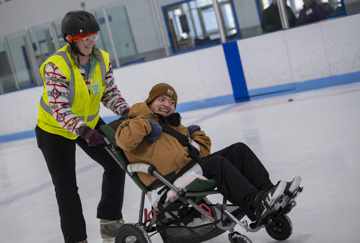 A man seated in a wheelchair is laughing while being pushed on the ice by AOA staff.  