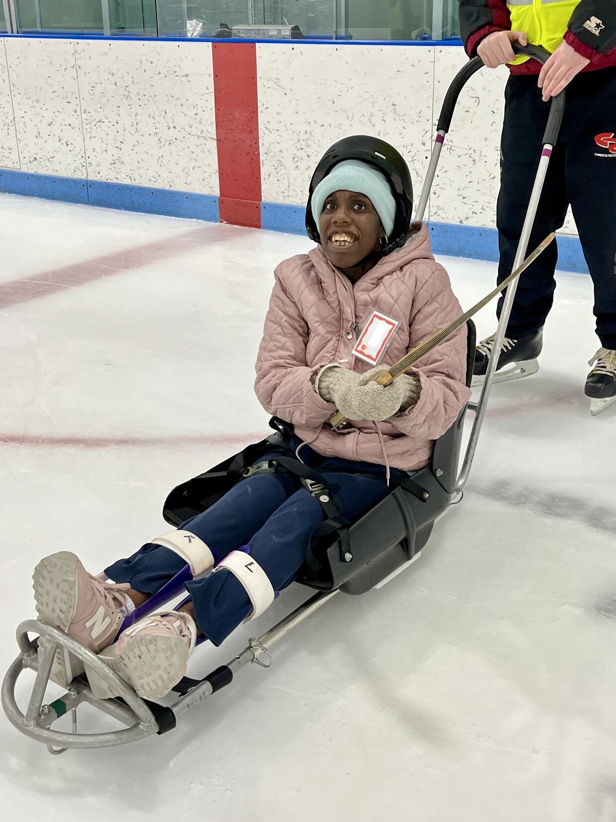 A young woman is smiling at the camera while seated in an ice sled holding a modified hockey stick.