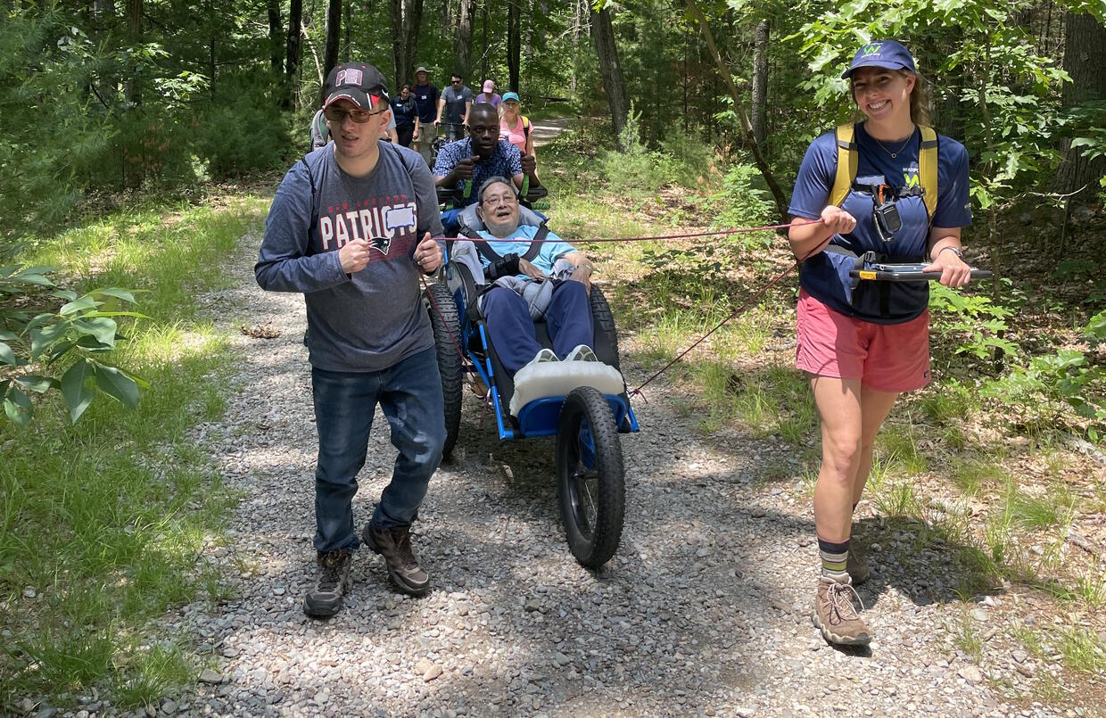 Hikers on a gentle trail in the woods. One hiker is using a three-wheeled recumbent wheelchair and being pulled with a rope.