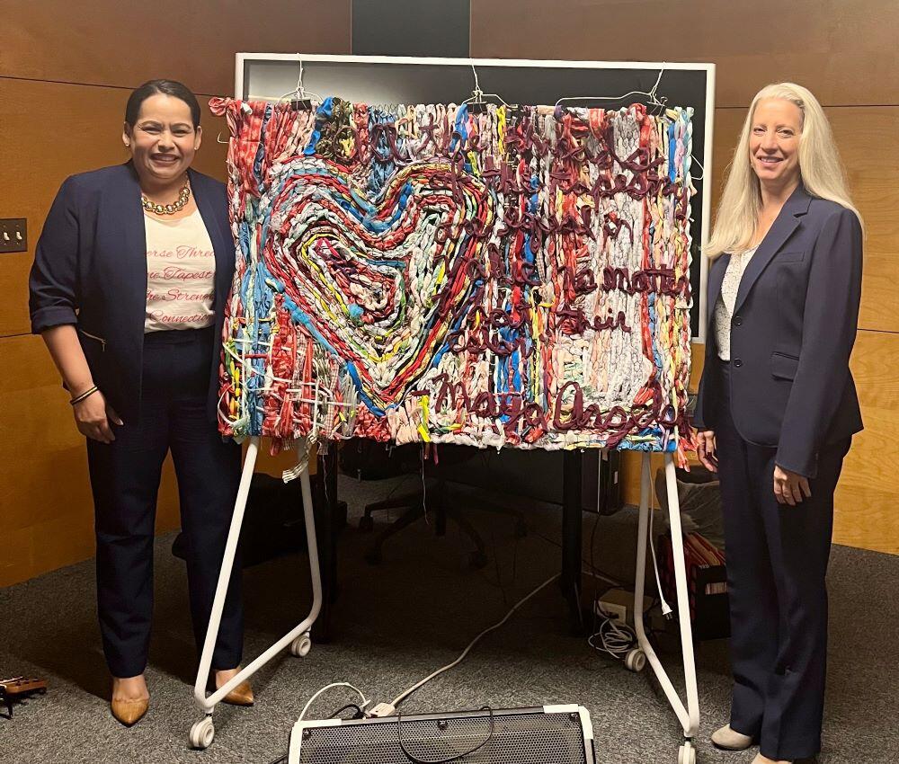 Two women stand on either side of a heart design tapestry. 