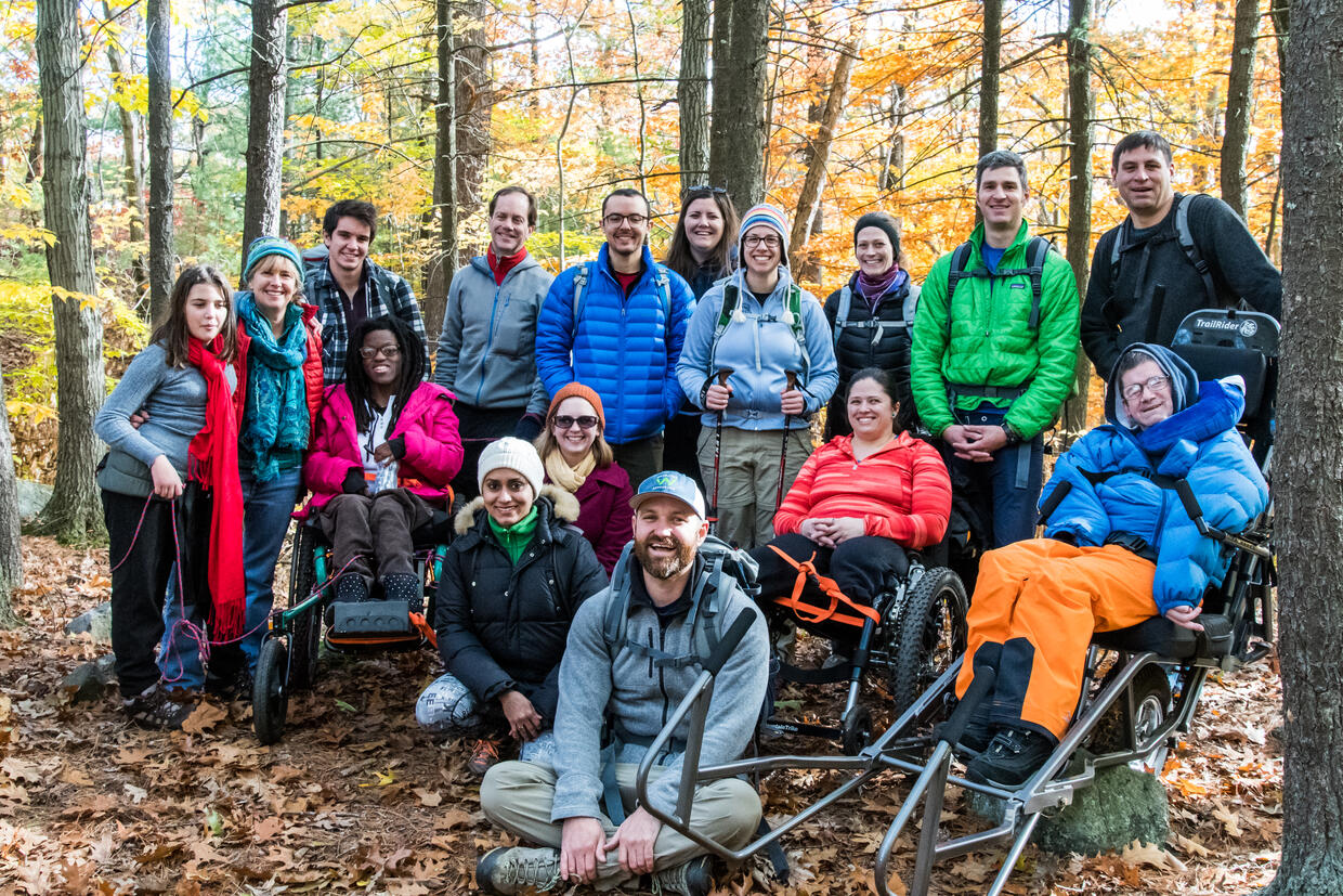 A group of hikers wearing winter gear are posing for the camera in a forest.  Some are seated in all-terrain wheelchairs and some are holding hiking sticks.