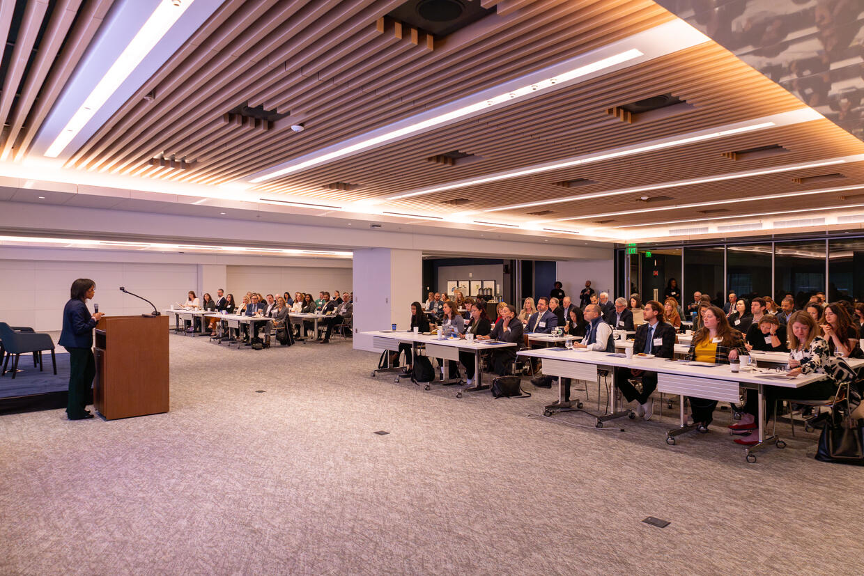 A picture taken from the corner of the room. Secretary Jones standing at a podium welcoming attendees to the convening. 