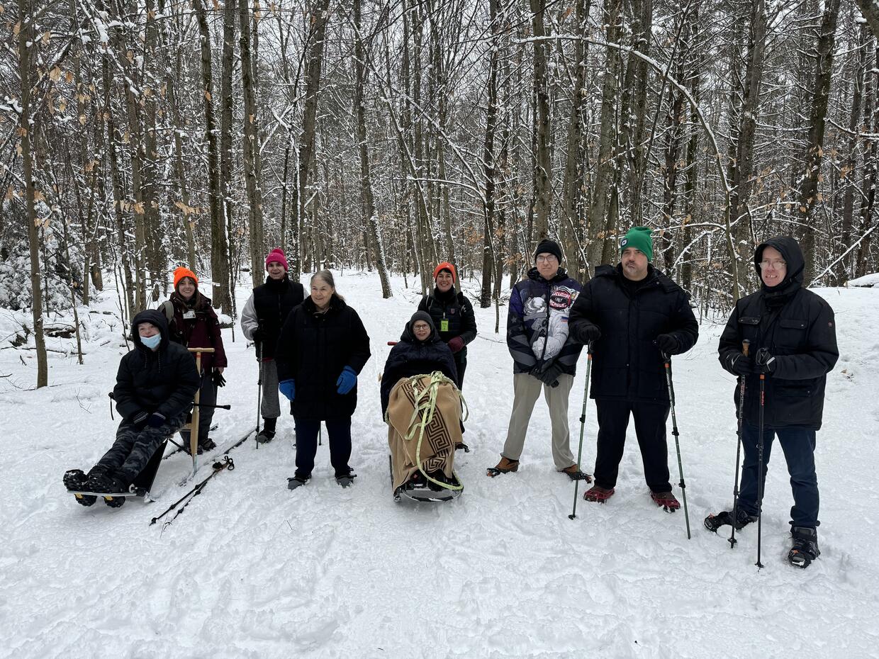 Group of hikers are smiling for the camera in a snowy forest. Some are  using snowshoes and poles, and some are seated on kick sleds.  