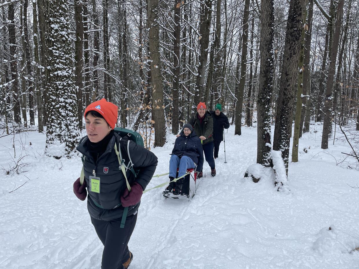 A person seated on a kicksled is being assisted along a snowy trail in the forest. One staff member is  pushing and one is pulling the kicksled. 