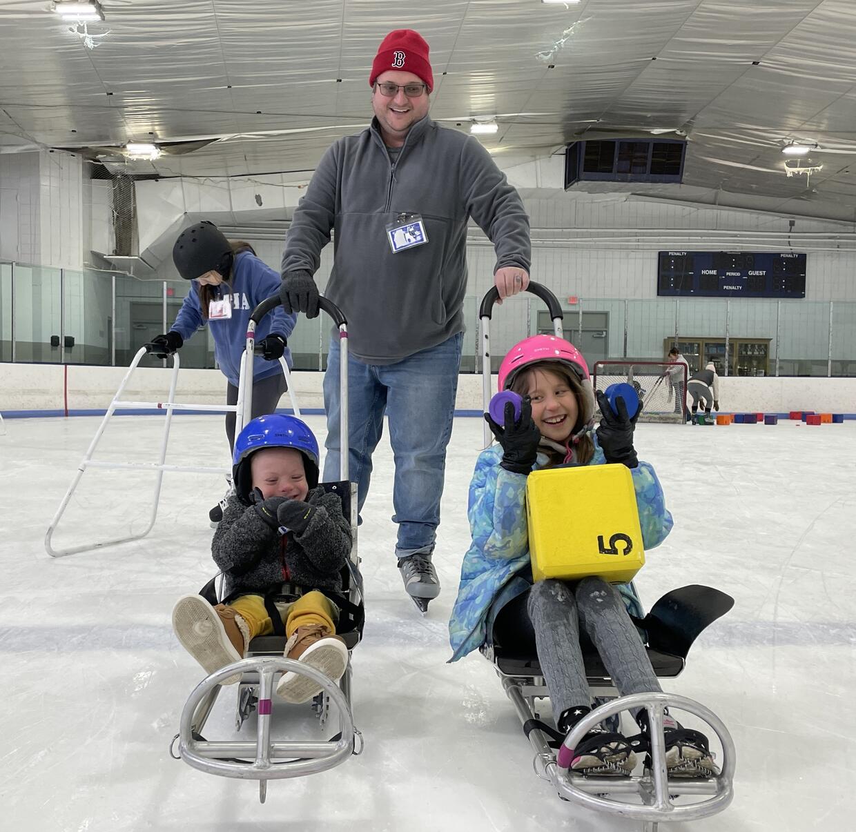 Two children seated in ice sleds are laughing while their father  stands behind them on the ice holding their sled handles. 