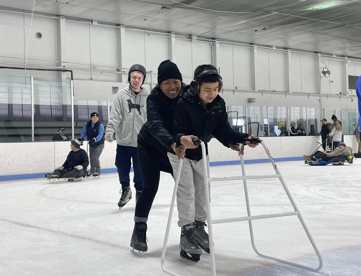 A young boy wearing headphones is skating with an ice walker. His parent is assisting from behind.  People skating with ice sleds are in the background. 