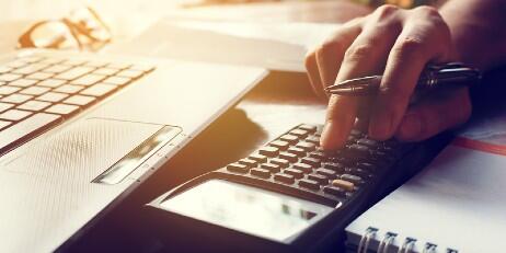 A hand holding a pen and using a calculator, next to a laptop