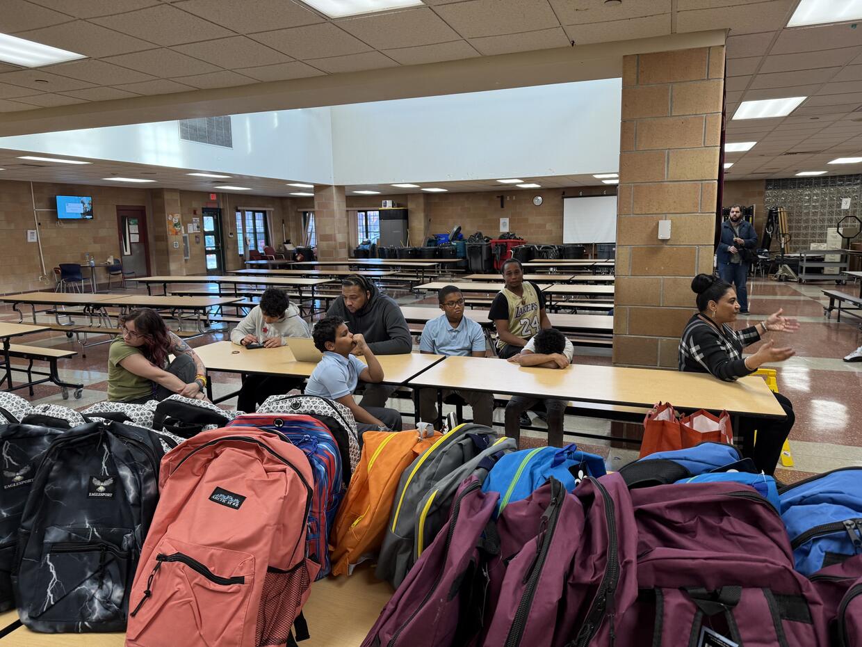 Cafeteria with people at a table and multi-colored backpacks in the foreground.