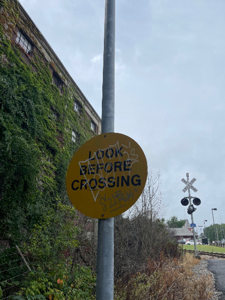 This image shows an obstructed “Look Before Crossing” sign at a train crossing at Walpole Station.