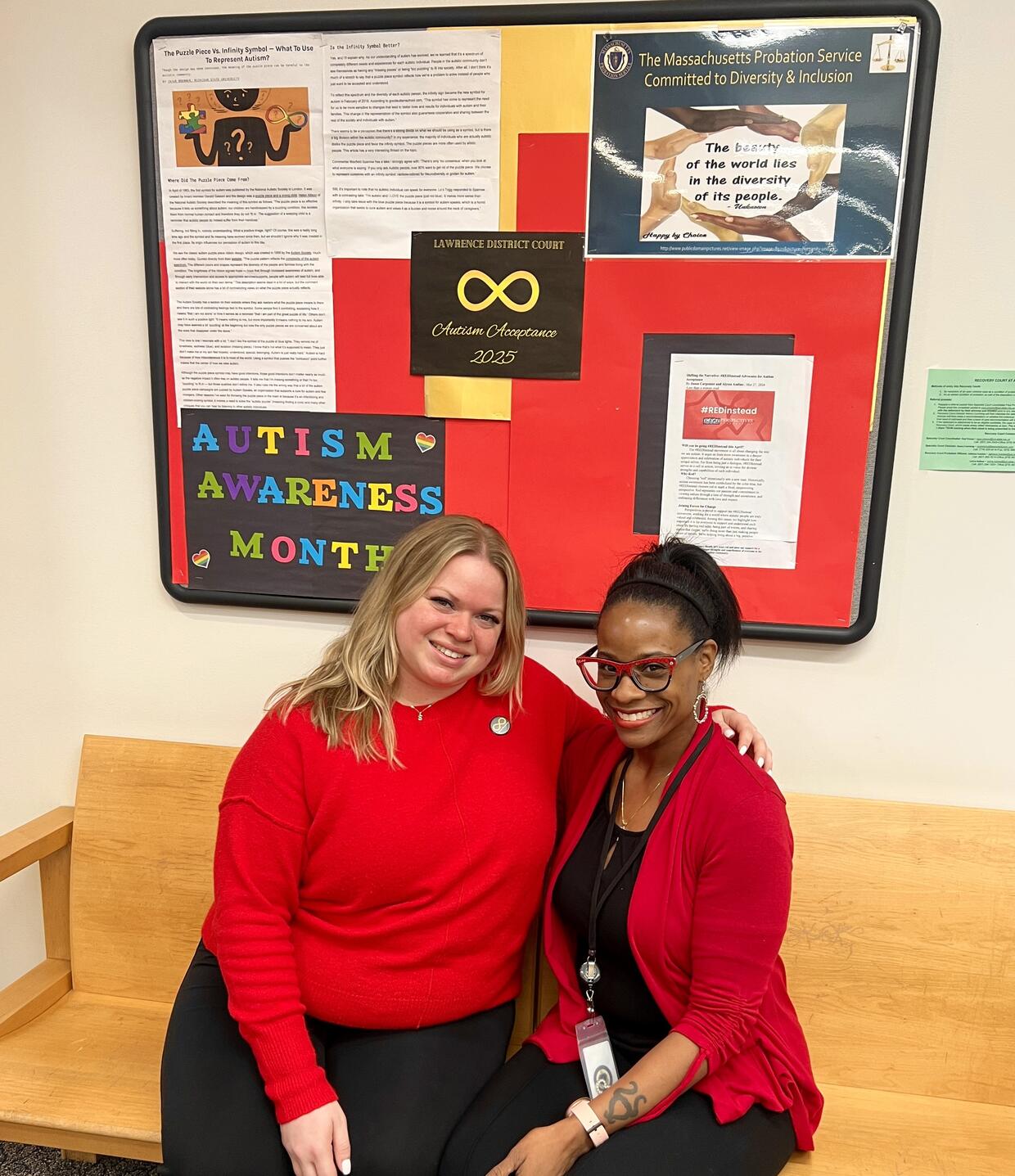 Two women  in red pose together under a push board decorated for Autism Awareness Month.
