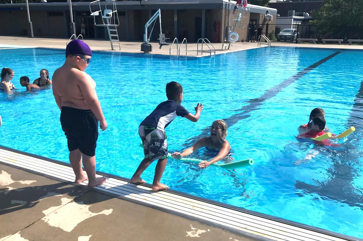 A child is jumping from the edge of the pool into the arms of staff holding a floatation noodle. 