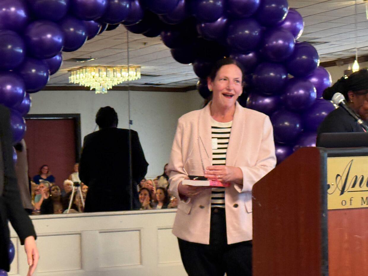 Person holding an award near a podium under a purple balloon arch in an event space.