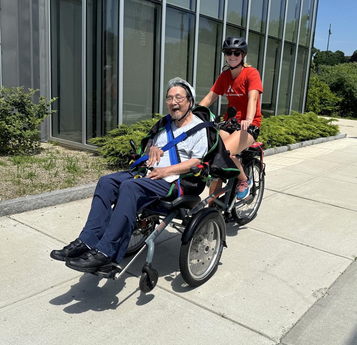 Two people riding a wheelchair tandem pose for a picture in front of a visitor center. 