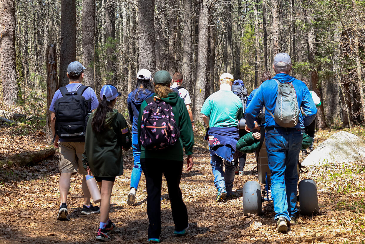 A group of people are hiking on a trail in the woods. One person is being pushed in an all-terrain wheelchair. 