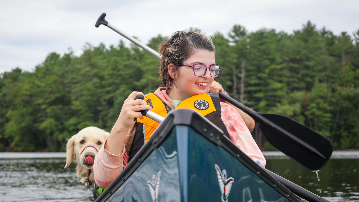 Young person paddling at the front of a canoe is smiling. A dog is seated in the middle. 