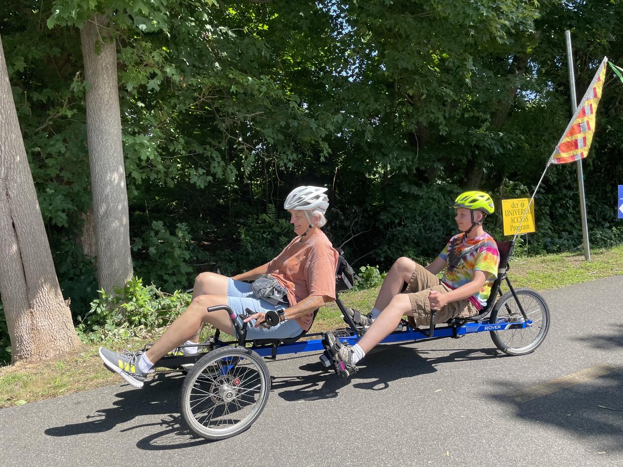 Two people are riding a recumbent tandem bike with a bright flag on a paved bike trail. 