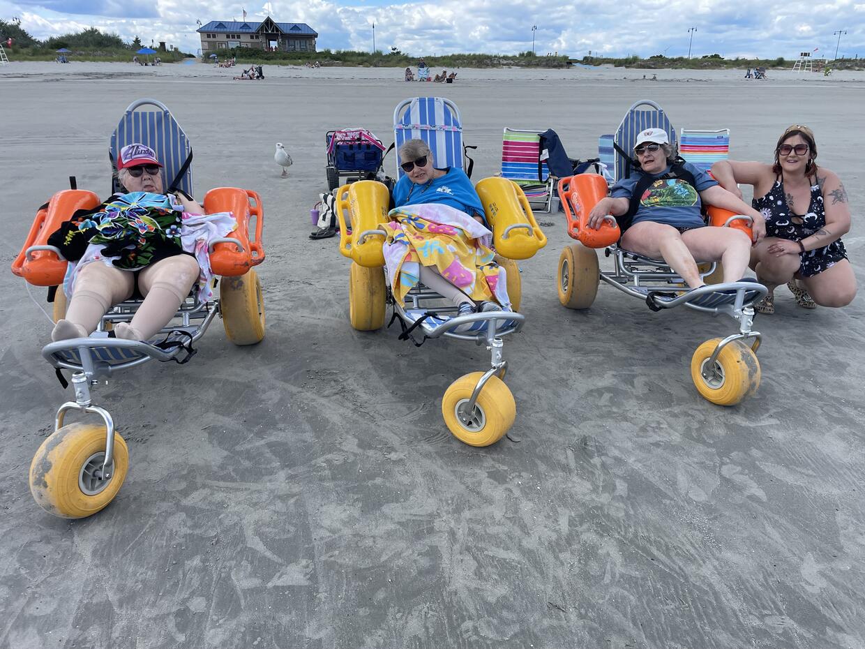 A group of people seated in floating beach wheelchairs on a sandy beach, smile for the camera. 