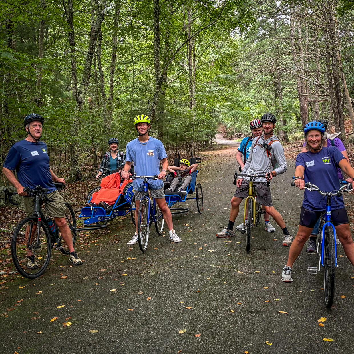 Group of people riding upright anda adaptive tandem smile for he camera on a paved trail in the woods.