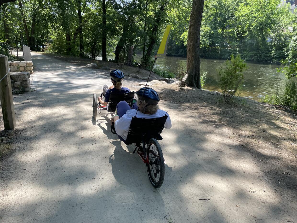 Two people are riding a recumbent tandem along a dirt trail next to a tree-lined river. 