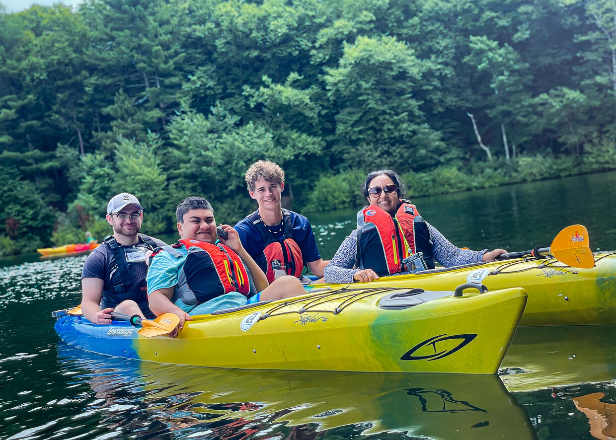 People seated  in two tandem kayaks on a tree-lined lake, smile for a photo. 