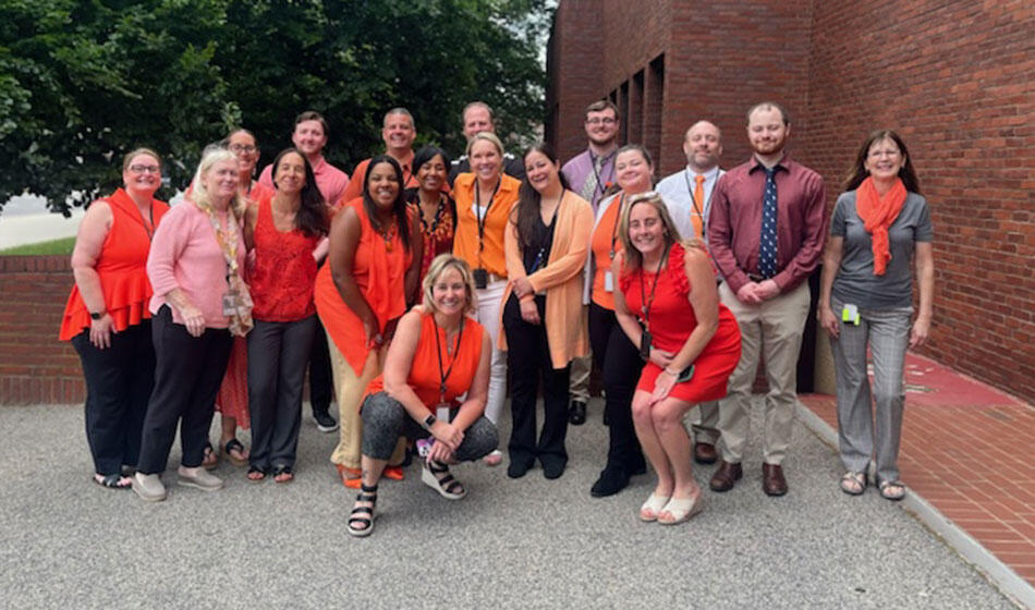 A group of nineteen people dressed in orange hues, posing outdoors near a brick building.