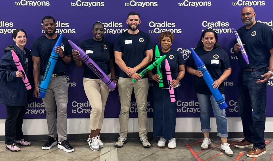A group of seven individuals stands in front of a "Cradles to Crayons" banner holding oversized crayons.