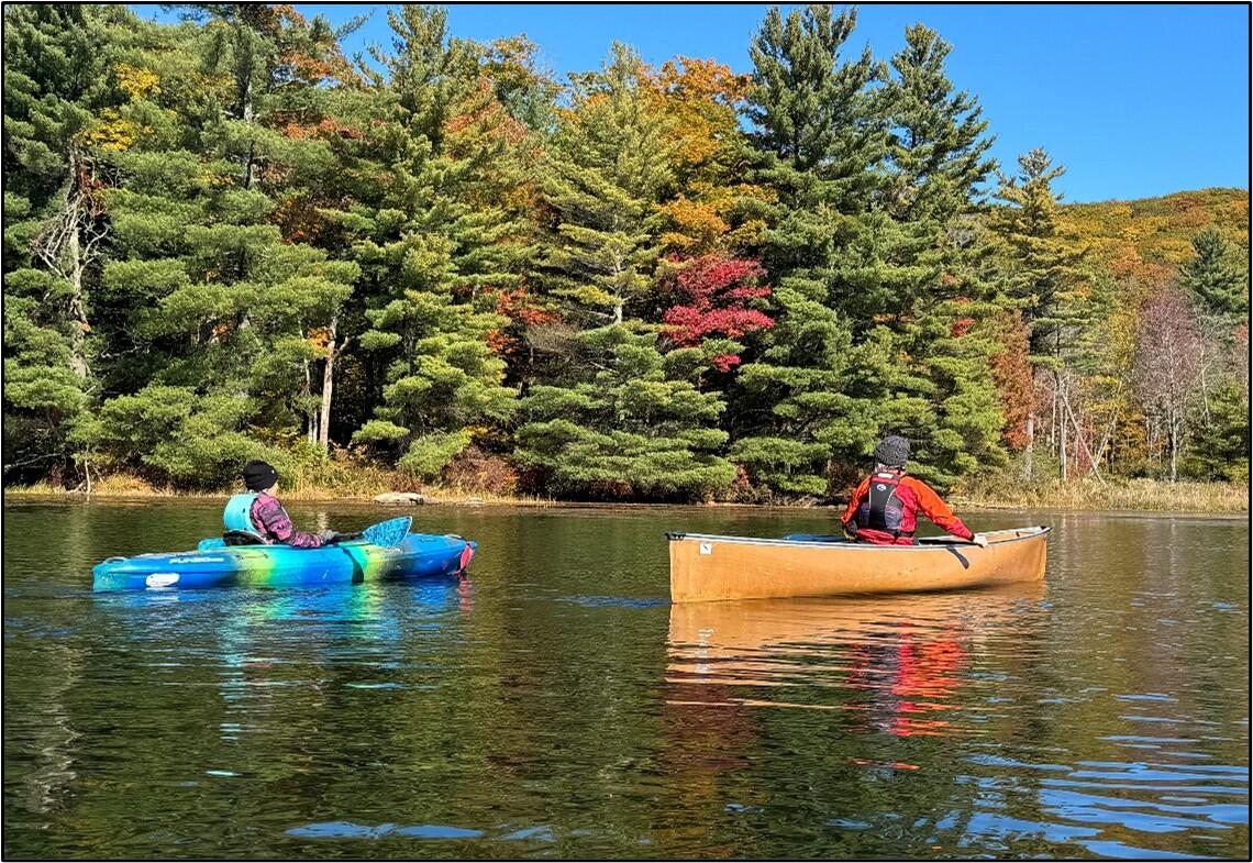 A kayaker and canoer are resting on a lake lined with pines and autumn trees. 