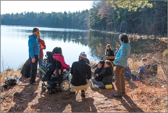 A group of hikers stop to talk along the edge of a pond in autumn. One person is seated in an all-terrain wheelchair. 