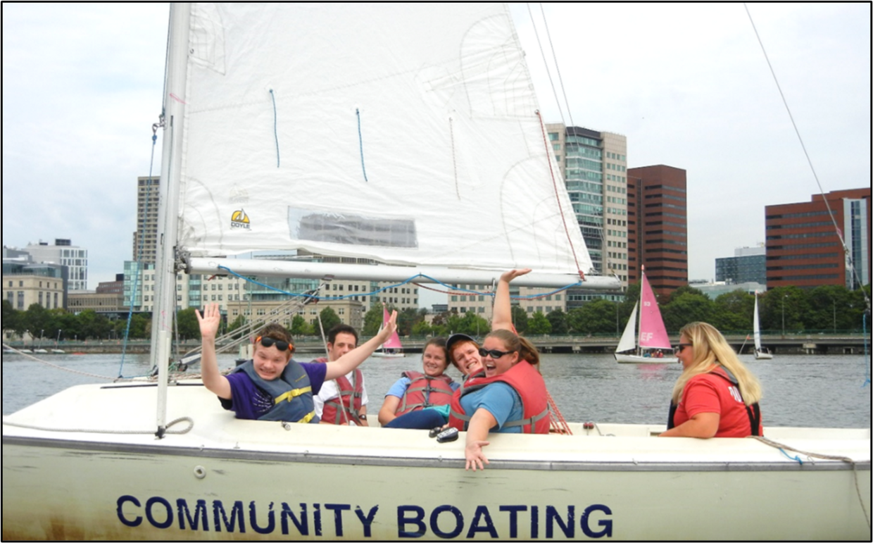 A group of people in a sailboat on the river are smiling and waving at the camera. 