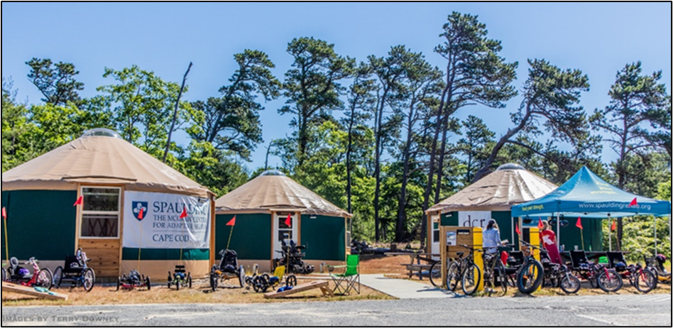 Many styles and sizes of adaptive bicycles are parked in front of 3 accessible yurts and a Spaulding pop up canopy.  