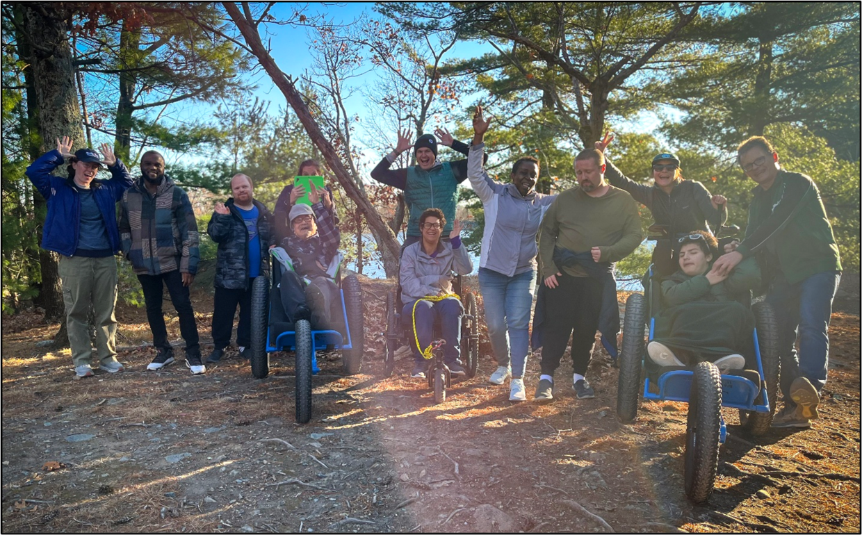 A group of hikers smile and wave on a dirt trail in the woods. Three are seated in all-terrain wheelchairs. 