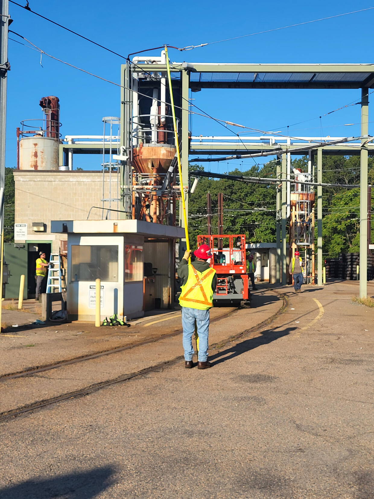 A worker in a yellow and orange safety vest uses a long yellow pole to test overhead power lines.