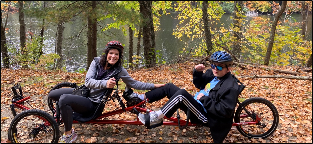 Two people seated on a recumbent tandem cycle, smile for a photo an a trail along a river in autumn.