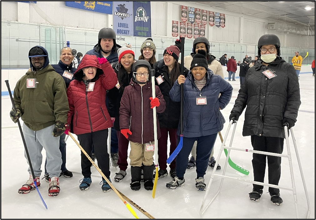 A group of people wearing Yaktrax smile for a photo on the ice.  Some are holding hockey sticks and one is using an skate walker. 