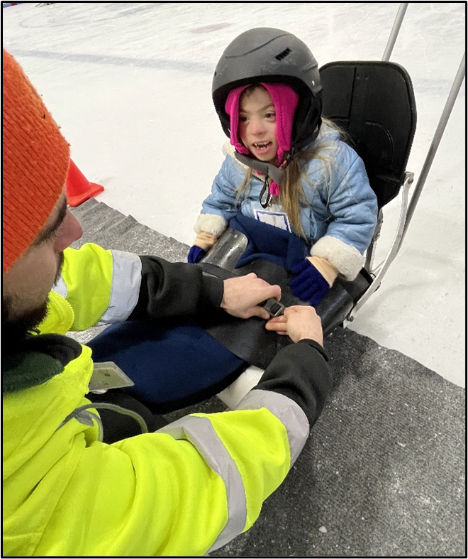 A young person seated on an ice sled smiles while UAP staff tie down a  lap blanket.