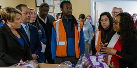 Governor Maura Healey and a group of officials and workers in orange vests listen attentively to a woman speaking near a table with packaged goods and supplies.