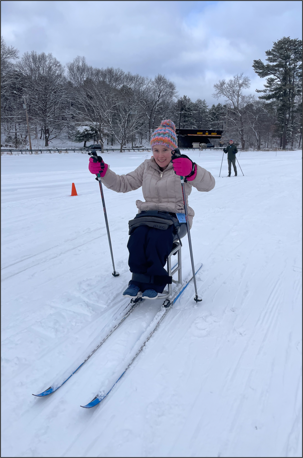 A young adult smiles while pushing themselves across the snow on a sit ski with ski poles.   